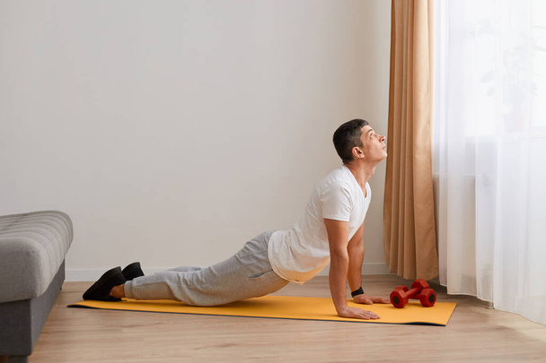 Full length portrait of sporty man practicing advanced yoga, yoga pose, stretching his body, wearing white t shirt and gray pants, fitness at home on yoga mat. - Photo, image