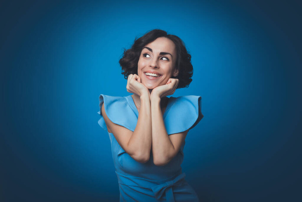 Close up portrait of an excited happy gorgeous girl in a long blue dress, who is looking in the camera while she posing with bright toothy smile and isolated on blue background - Foto, imagen