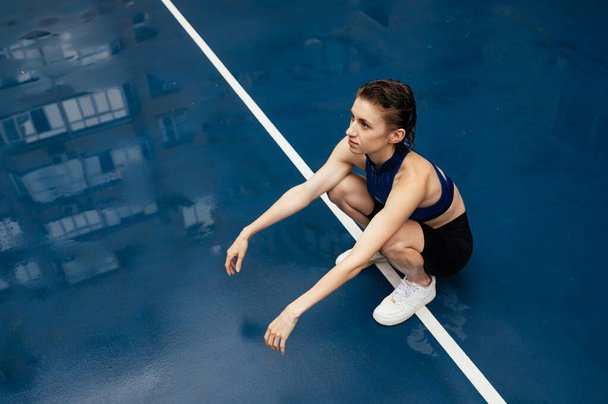 Atletische dame zit op een blauw sportveld en rust na de training, wegkijkend met een serieus gezicht. - Foto, afbeelding