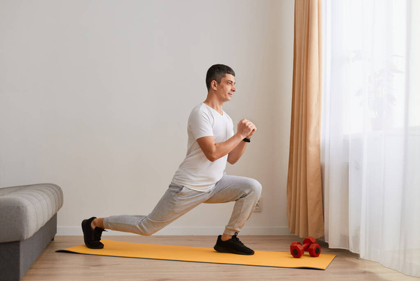 Indoor shot of athletic sporty young man with dark hair wearing white t shirt and gray pants, doing sport lunge exercise, warming up training muscles, butts workout at home. - Zdjęcie, obraz