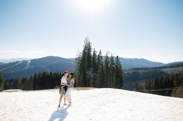 Νεαρό κομψό ζευγάρι στέκεται στο χιόνι στα βουνά αγκαλιάζει σε μια ηλιόλουστη μέρα του καλοκαιριού και χαμογελώντας με φόντο το όμορφο ορεινό τοπίο. - Φωτογραφία, εικόνα