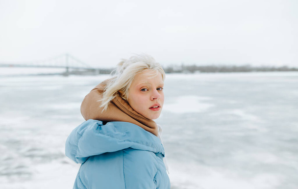 Portret pięknej dziewczyny z blond włosami i brwiami na zimowej, śnieżnej ulicy na tle zamarzniętego jeziora, patrzącej w kamerę. - Zdjęcie, obraz