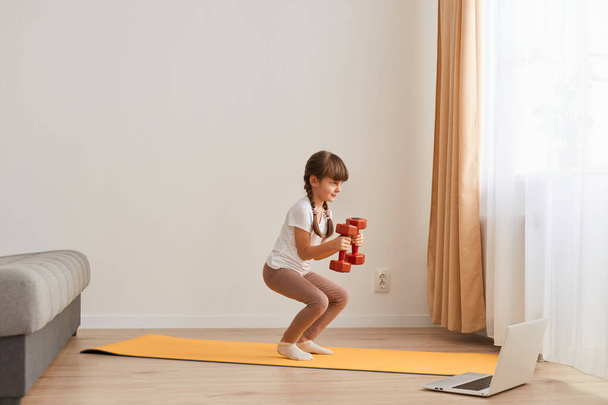 Portrait of female kid doing squatting workout at home, cute kid training on a mat indoor, dark-haired female kid having exercises with dumbbells in hands near the window. - Photo, image