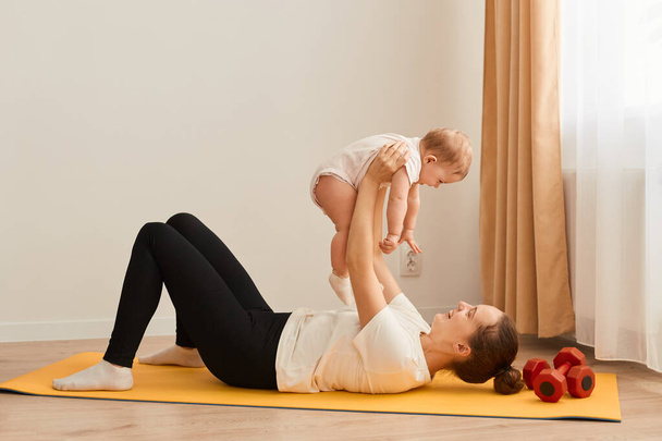 Indoor shot of of athletic woman wearing white t shirt and black leggins lying on yoga mat on floor, doing sport exercises with her baby daughter in hands, expressing positive emotions. - Фото, зображення