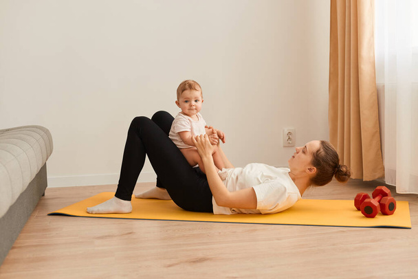 Horizontal shot of mother and baby daughter doing gymnastics, female practicing yoga exercises on floor on yoga mat at home, fitness and healthy lifestyle. - Fotoğraf, Görsel