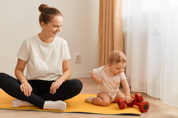 Indoor shot of athletic woman wearing white t shirt and black leggins sitting on floor on yoga mat with her toddler daughter, looking at baby with happy smile, fitness at home. - Foto, afbeelding