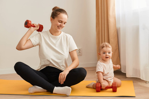 Indoor shot of sporty athletic woman in white t shirt and black leggins sitting on yoga mat and training arms with raising dumbbell, doing sport exercises with her infant daughter. - Foto, afbeelding