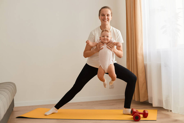 Indoor shot of sporty young adult mother doing lunges and holding toddler daughter in hands, fitness at home while looking after little child, healthy lifestyle. - Foto, Imagem