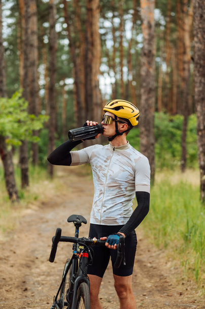 Male cyclist stands with a bicycle in the woods on a background of beautiful natural views and drinks water from a bottle. - Zdjęcie, obraz