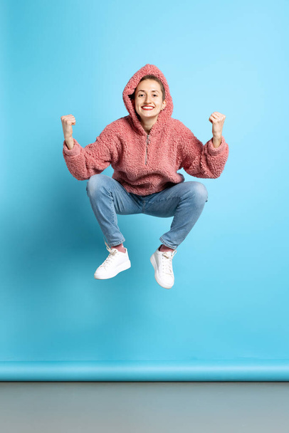 Dynamic portrait of cool young girl, student with short hair wearing pink hoodie jumping isolated on blue background. Concept of emotions, music - Fotografie, Obrázek