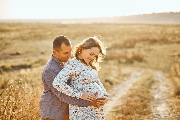 husband with pregnant wife with big belly tummy is resting on bank of river in summer sunny weather. Happy couple in love caucasian man and woman hugging outdoors - Photo, image