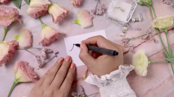 A kezek RSVP-t írnak egy kártyára, közel a rózsaszín virágokhoz, közel a márványasztalhoz. - Felvétel, videó