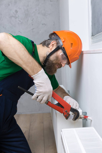 Ein männlicher Klempner installiert einen Heizkörper in der Heizungsanlage einer Wohnung. Kerl in Overalls und einem Gasschlüssel. - Foto, Bild