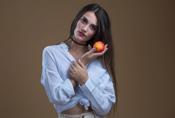 Studio portrait of a beautiful girl in a white shirt with a peach on a beige background - Фото, зображення