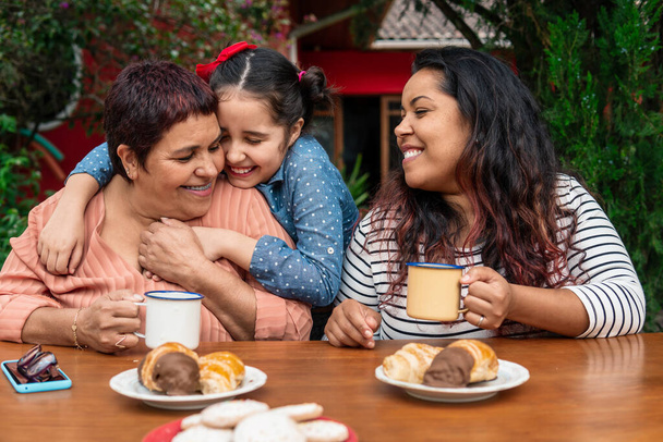 Βραζιλιάνα εγγονή αγκαλιά γιαγιά κατά τη διάρκεια του πρωινού στο τραπέζι έξω - Φωτογραφία, εικόνα