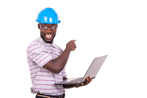 jovem engenheiro vestindo capacete de segurança azul, segurando computador portátil e apontando o dedo para quadro branco enquanto sorri. - Foto, Imagem