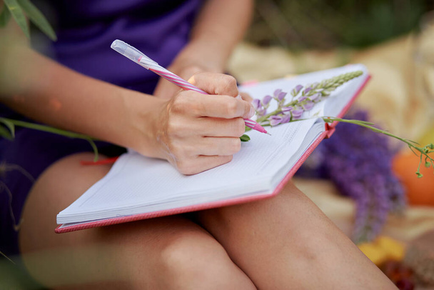Kobieta pisząca w notatniku z bukietem dzikich kwiatów - łubin. Bliskość natury, koncepcja samopoznania. Zamknij drzwi. Relaks i dobre samopoczucie koncepcja. Słoneczny dzień, dobry nastrój. - Zdjęcie, obraz