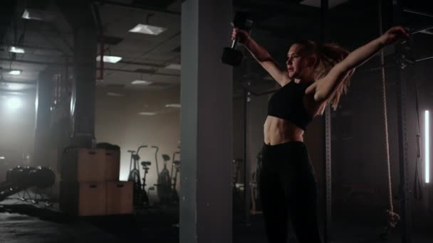 Σε αργή κίνηση, μια γυναίκα σηκώνει ένα βαράκι πάνω από το κεφάλι της ενώ γυμνάζεται σε ένα σκοτεινό γυμναστήριο. Αθλητική ισχυρή γυναίκα εκτελεί μια δύσκολη προπόνηση με την ανύψωση αλτήρες - Πλάνα, βίντεο