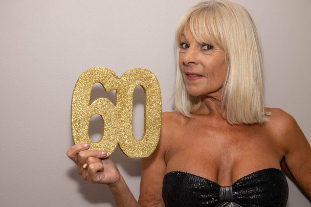 Una modelo femenina mayor madura con un vestido negro con la mujer de pelo rubio sosteniendo un cartel que dice 60, 60º concepto de celebraciones de cumpleaños - Foto, Imagen