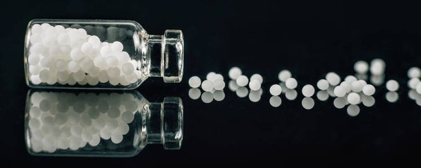 Гомеопатия - Гомеопатические глобулы, разбросанные из стеклянной бутылки на черном фоне - Фото, изображение