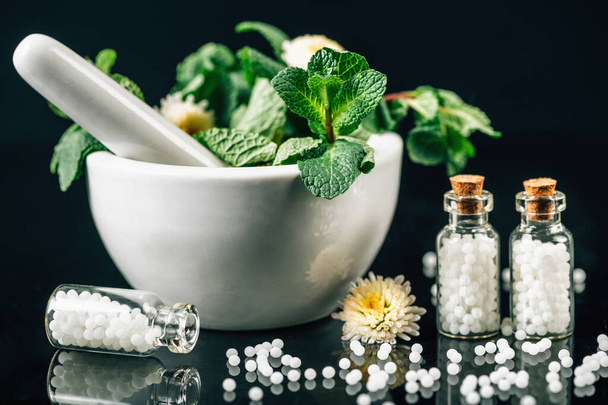 Homeopatia Tratamento - glóbulos homeopáticos espalhados para fora do frasco de vidro, argamassa e pilão cheio de plantas medicinais frescas, fundo preto - Foto, Imagem