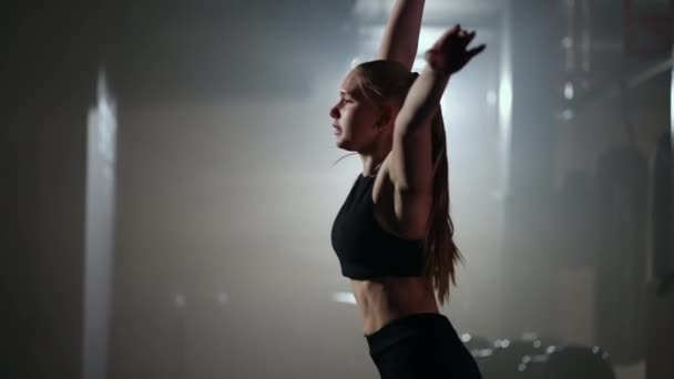 Lassított felvételen egy nő felemel egy súlyzót a feje fölé, miközben egy sötét edzőteremben edz. Atlétikai erős nő végez egy nehéz edzés emelő súlyzók - Felvétel, videó