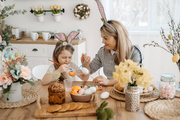 Cute little girl in bunny ears with her mother paint eggs while sitting at the table in the kitchen. Decorations for the celebration of Easter - Foto, Imagen