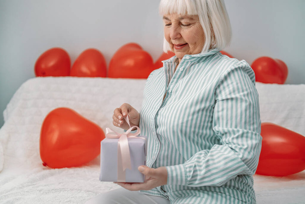Mujer rubia de 50 años en una elegante blusa desempaqueta una caja de regalo sorpresa el 14 de febrero sentada en un sofá en casa. - Foto, imagen