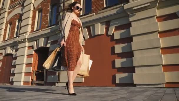 Attractive young lady holds shopping bags walking at sidewalk after purchases. Fashionable woman carries paper packages going along urban street. Concept of leisure and free time. Low view Slow motion - Footage, Video
