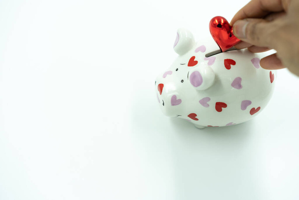 Hand from mixed race woman fills white piggy bank with red heart on white background. - Photo, Image