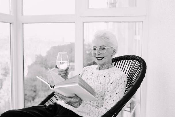 Terasta oturan eski moda son sınıf tarzı bir kadın. Elinde beyaz şarap kadehiyle kitap okuyor.. - Fotoğraf, Görsel