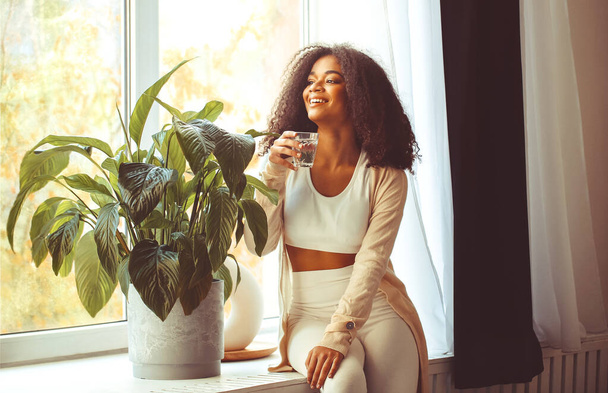 Jeune femme afro-américaine positive assise sur le rebord de la fenêtre avec un verre d'eau minérale pure et regardant par la fenêtre avec le sourire, buvant de l'aqua le matin, commençant une nouvelle journée avec de saines habitudes - Photo, image