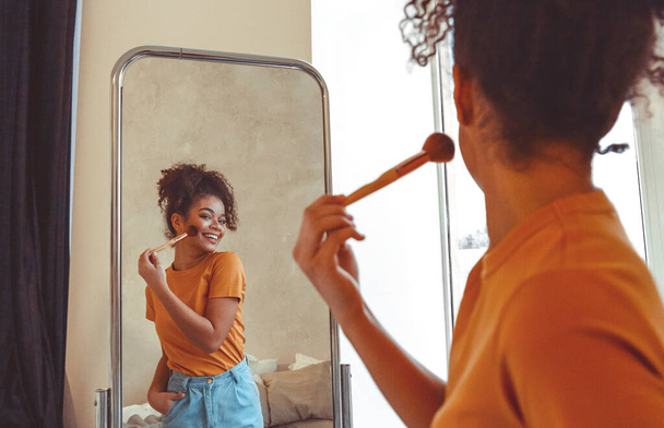 Χαρούμενη χαρούμενη Αφρο-Αμερικανίδα νεαρή γυναίκα που βάζει μακιγιάζ με πινέλο, στέκεται μπροστά από έναν μεγάλο καθρέφτη στο σπίτι, κοιτάζοντας τη δική της αντανάκλαση με πλατύ χαμόγελο. Έννοια ρουτίνας ομορφιάς - Φωτογραφία, εικόνα