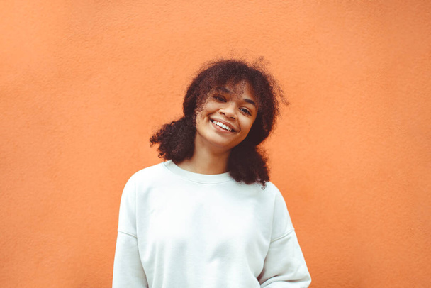 Καθαρή ευτυχία. Χαριτωμένο χαρούμενο αφροαμερικανό κορίτσι με σγουρό χτένισμα που φοράει λευκό πουλόβερ και ποζάρει στον πορτοκαλί τοίχο. Χαμογελαστή μαύρη γυναίκα που νιώθει ικανοποιημένη με τη ζωή της. - Φωτογραφία, εικόνα