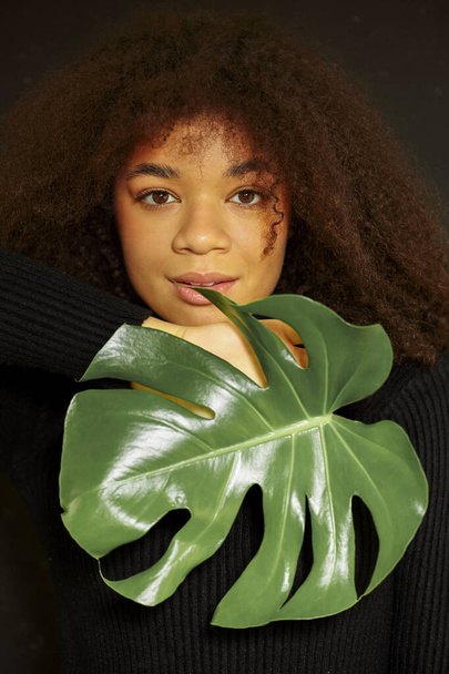 Φυσική ομορφιά. Σκούρο πορτρέτο στούντιο της νεαρής αφροαμερικανής γυναίκας με πράσινα φύλλα monstera και κοιτάζοντας κάμερα, σκεπτική αφρικανική θηλυκό εκμετάλλευση τροπικό φυτό απομονωμένο πάνω από το μαύρο τοίχο - Φωτογραφία, εικόνα