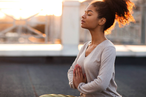 Foto recortada de una mujer sentada en loto posando sobre una esterilla y practicando yoga, una mujer haciendo gesto namaste mientras medita en el interior. Concepto de meditación y atención plena - Foto, imagen
