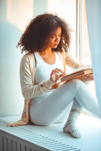 Mein Hobby ist das Lesen. Ruhige entspannte afrikanisch-amerikanische junge Frau liest Buch, während sie zu Hause auf der Fensterbank sitzt, schwarz gelocktes Mädchen am Fenster entspannt die Freizeit am Wochenende verbringt, vertikale Aufnahme - Foto, Bild