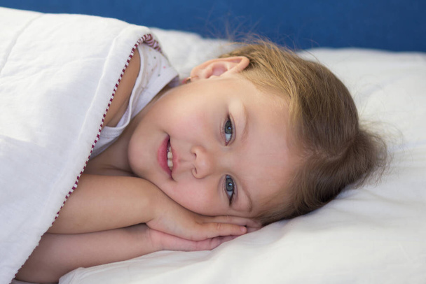 Valkoihoinen hymyilevä kolmevuotias lapsi makaa sängyssä käsi pään alla yrittäen nukkua päivän aikana. - Valokuva, kuva