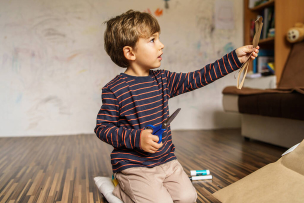 Ritratto frontale del bambino di quattro anni seduto sul pavimento a casa da solo con le forbici che guardano a lato - concetto di sviluppo dell'infanzia e tempo libero - Foto, immagini