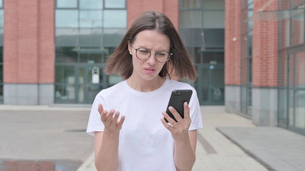 Young Woman Reacting to Loss on Smartphone while Walking on Street - Foto, Imagen