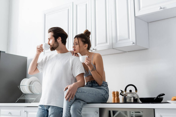 young woman with tattoo holding cup and sitting near boyfriend drinking coffee in kitchen  - Foto, Imagen