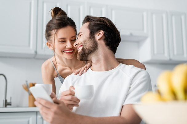 happy bearded man using smartphone near smiling woman in bra holding cup  - Valokuva, kuva