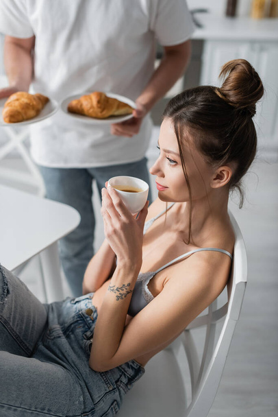 tattooed woman holding cup near blurred man with croissants  - Foto, imagen