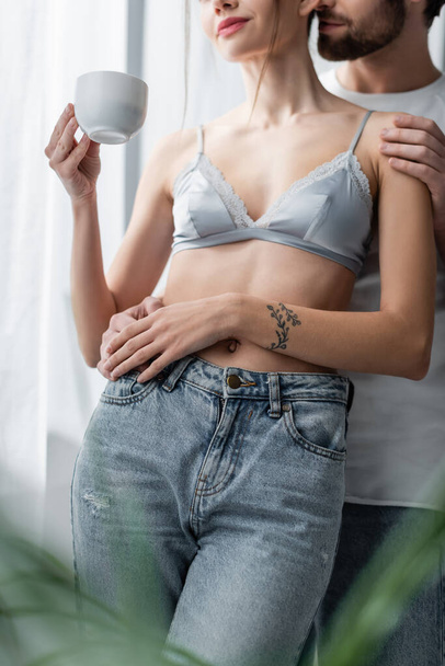 cropped view of man hugging tattooed woman in bra and jeans with cup  - Foto, imagen