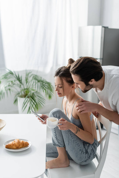 young woman in bra and jeans using smartphone and holding cup near boyfriend in kitchen  - Foto, Imagem