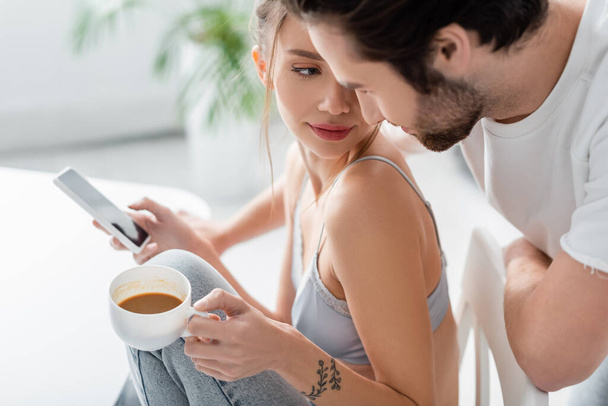 tattooed young woman in bra and jeans using smartphone and holding cup near boyfriend in kitchen  - Fotó, kép