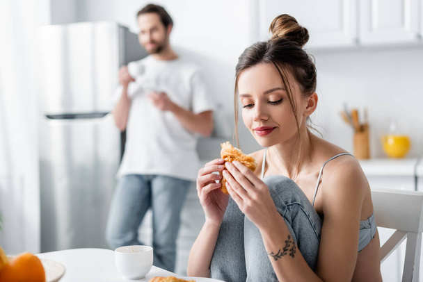 junge Frau isst Croissant neben verschwommenem Mann im Hintergrund  - Foto, Bild