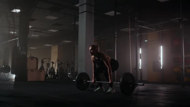 Slow motion: Een vrouw tilt een lange halter in een donkere sportschool met veel gewicht. Een sterke jonge vrouw tilt gewichten voor een training - Video