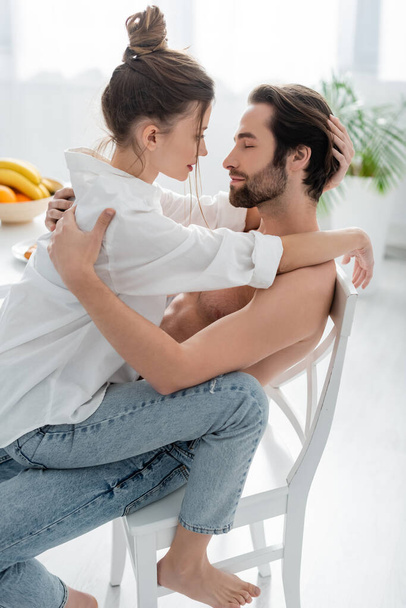 side view of passionate woman sitting on bearded and shirtless man in kitchen  - Photo, Image