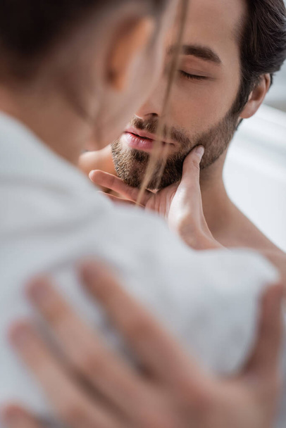 blurred woman touching face of bearded boyfriend with closed eyes  - Foto, afbeelding